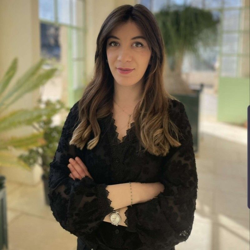 Une femme vêtue d'une chemise blanche et d'une salopette bleue sourit à la caméra