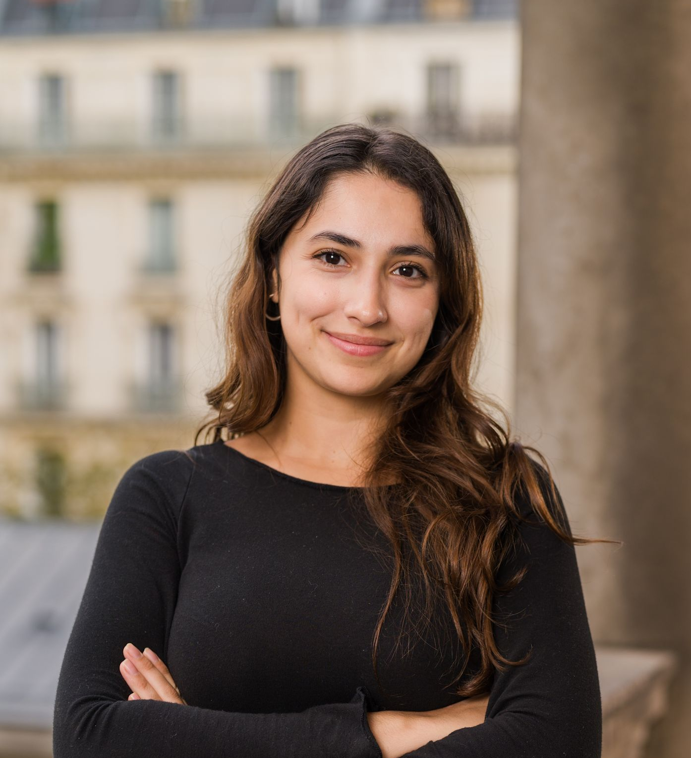 Une femme aux cheveux bouclés sourit à la caméra devant un immeuble.