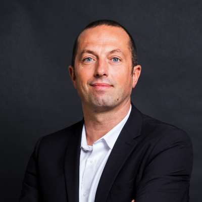 Un jeune homme avec une barbe et des lunettes de soleil sourit à la caméra.