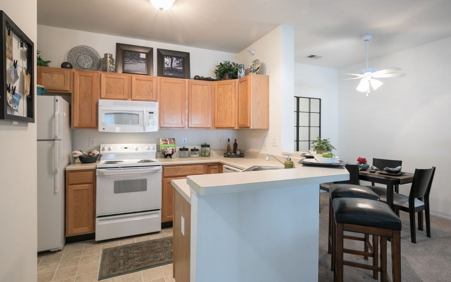 A kitchen with wooden cabinets, white appliances, and a breakfast bar. A dining area is visible in the background.