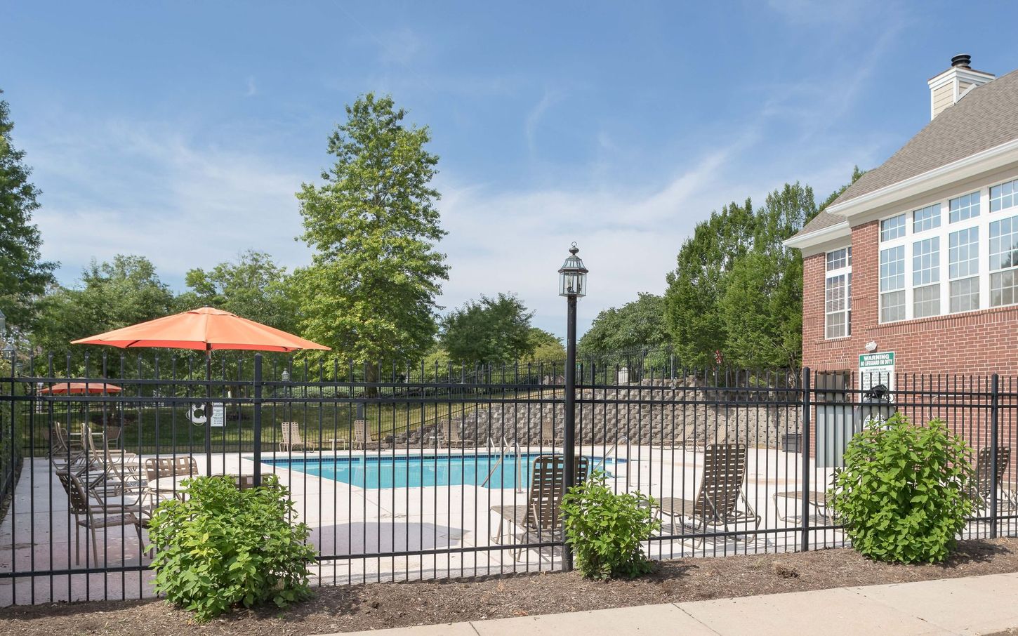 A fenced-in outdoor pool with an orange umbrella and lounge chairs. Next to a brick building under a blue sky.