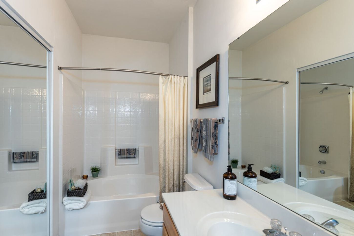 Bathroom with white tiles, tub, toilet, and vanity. A large mirror and a framed artwork are on the wall.