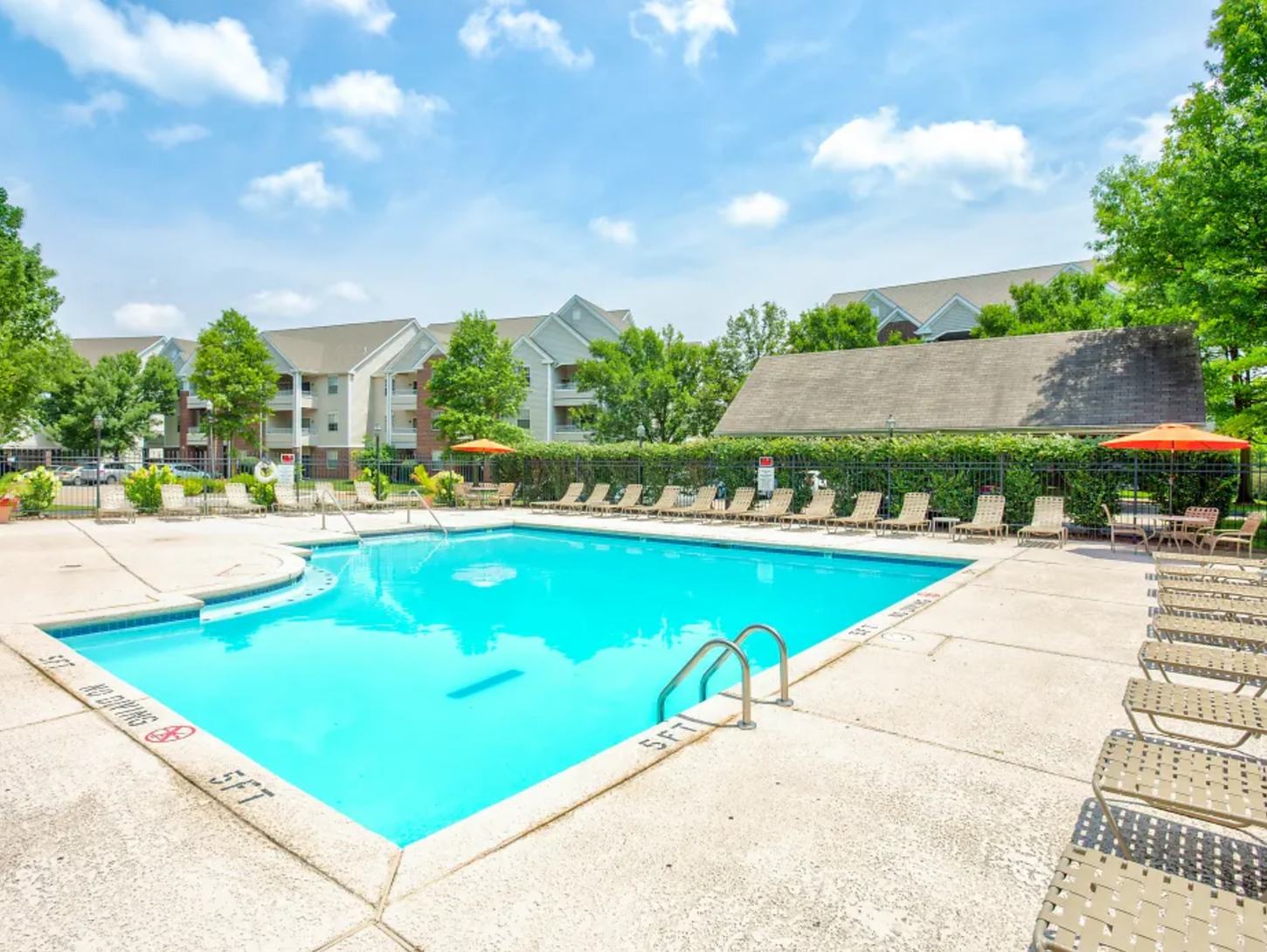 Swimming pool surrounded by lounge chairs and trees. Apartments are visible in the background under a blue sky.