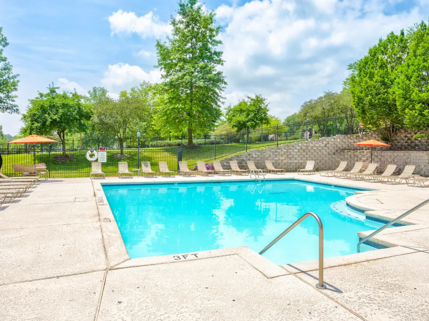 Outdoor swimming pool with clear blue water and lounge chairs. Green trees surround the pool under a sunny, blue sky.