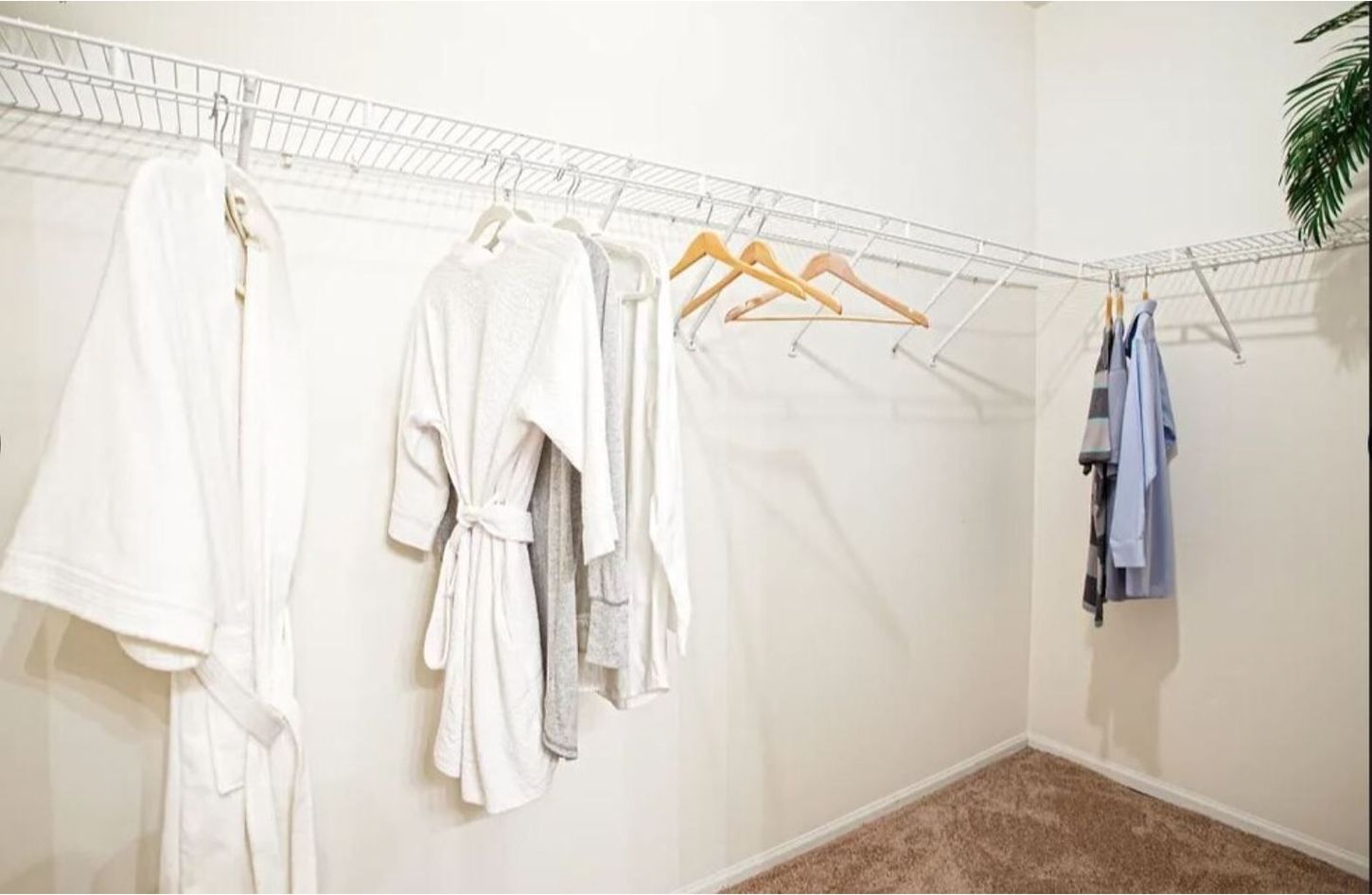 A white walk-in closet with hanging clothes, mostly white robes and a blue shirt, against neutral walls and carpet.