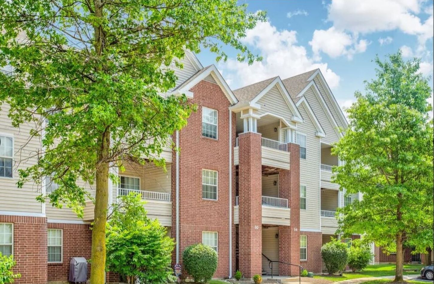 Apartment building with brick and beige siding, balconies, and green trees under a partly cloudy sky.