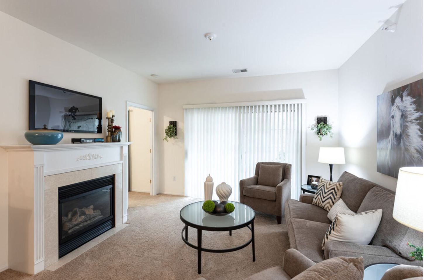 A living room with fireplace, TV, couch, armchair, coffee table, and sliding glass door to a patio. Beige walls and carpeting.