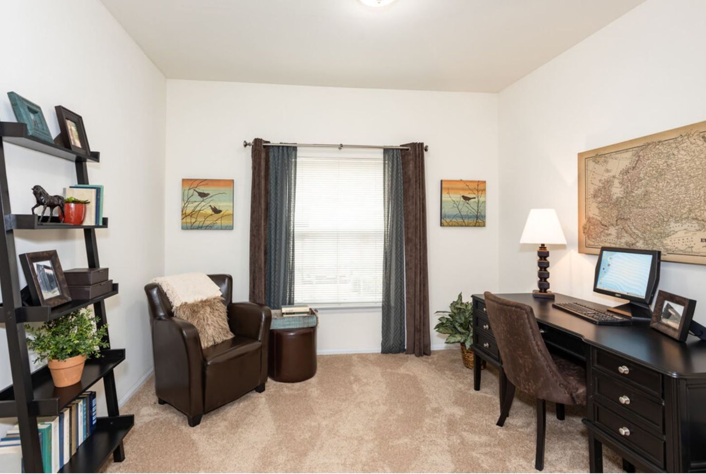 Home office with a dark wooden desk, chair, and bookshelf. A brown leather armchair sits near a window with brown and gray curtains; beige carpet.