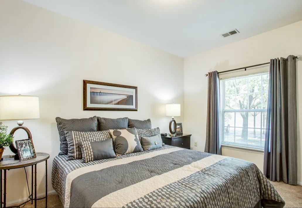 Bedroom with neutral walls, bed with gray bedding, two bedside tables, and a window with gray curtains.