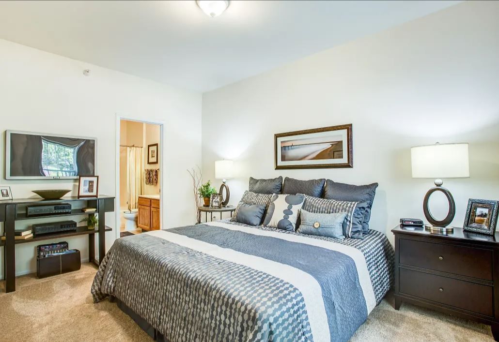 Bedroom with a bed, nightstands, a television, and a doorway to a bathroom. Neutral color scheme with dark brown furniture and a gray and white bedspread.