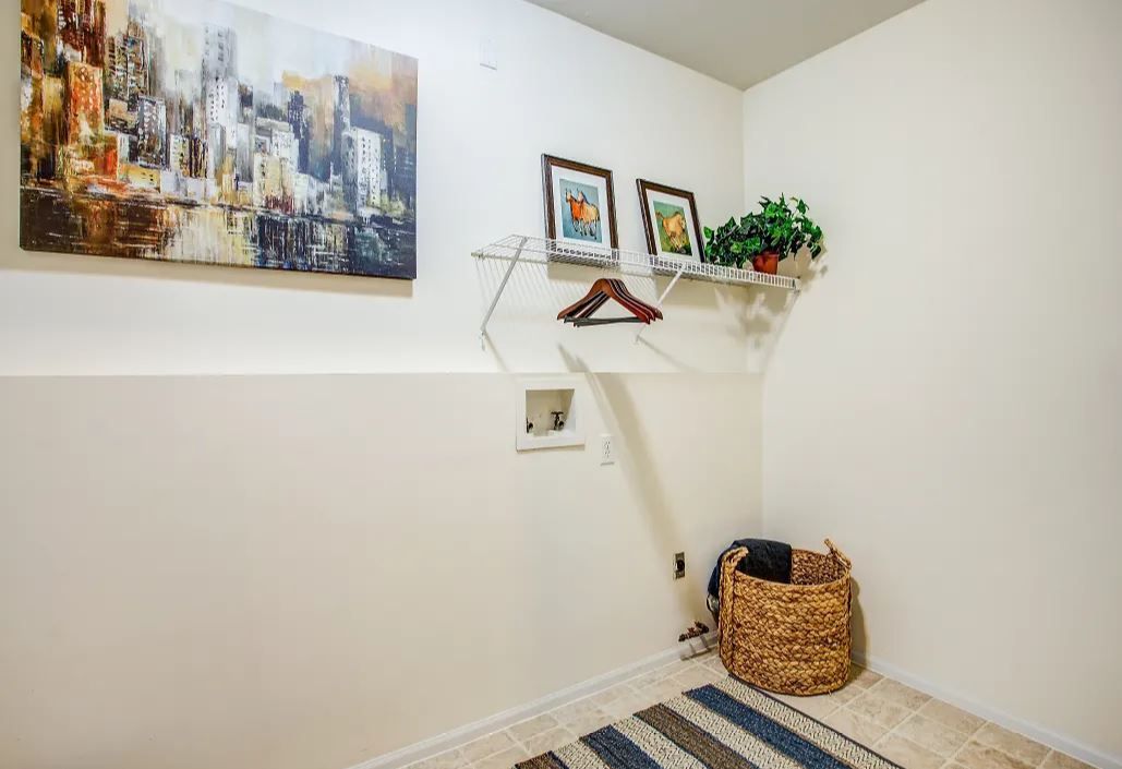 Laundry room with beige walls and tile, a painting, shelf with art and plant, clothes hamper, and rug.