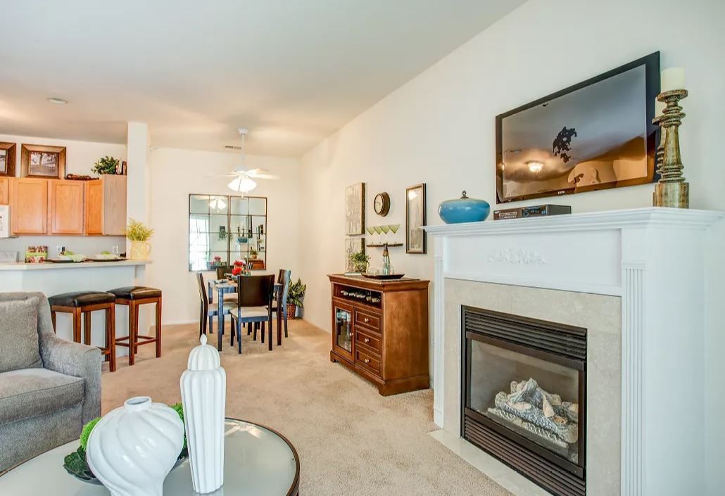 Living room with fireplace, TV, dining area, and open kitchen. Neutral colors, cozy furniture, and decorative items.