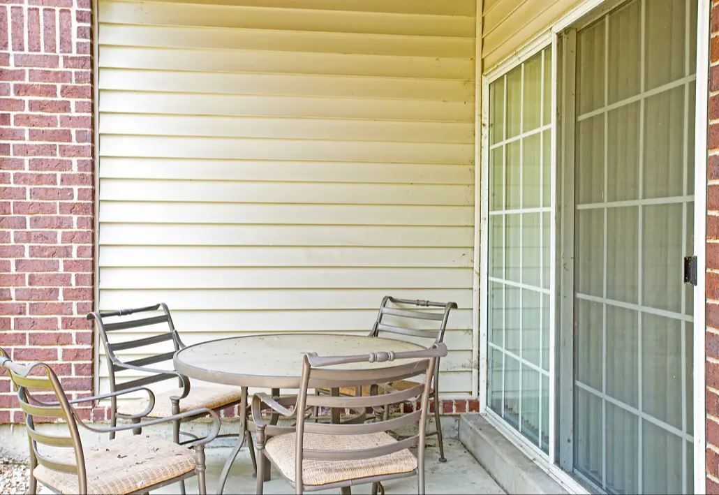 Outdoor patio with round table and four chairs, against a beige siding wall and a brick pillar, with a glass sliding door to the right.