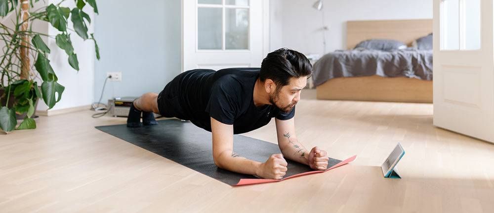 A man is doing a plank on a yoga mat in front of a tablet.