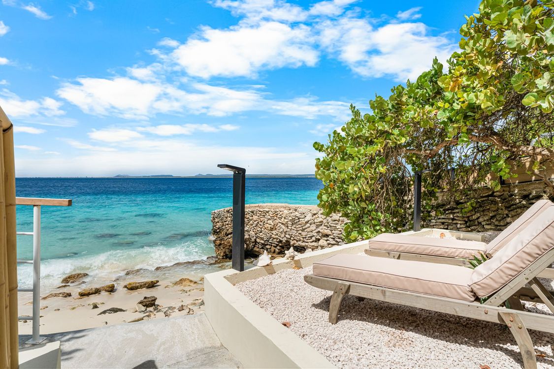 Two lounge chairs on a white pebbled patio overlooking a turquoise ocean under a blue sky.