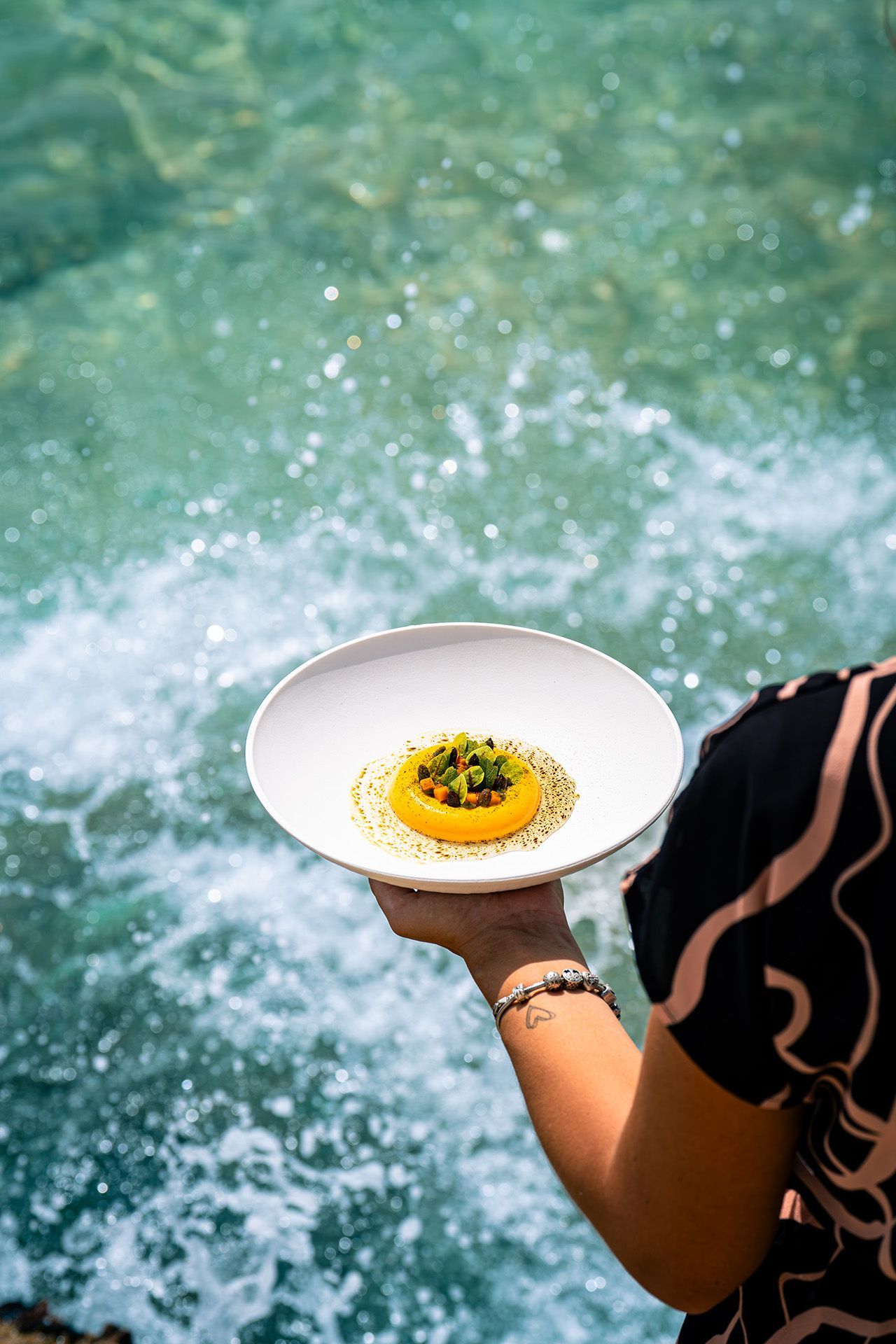 Person holding a plate with yellow food, ocean in the background.
