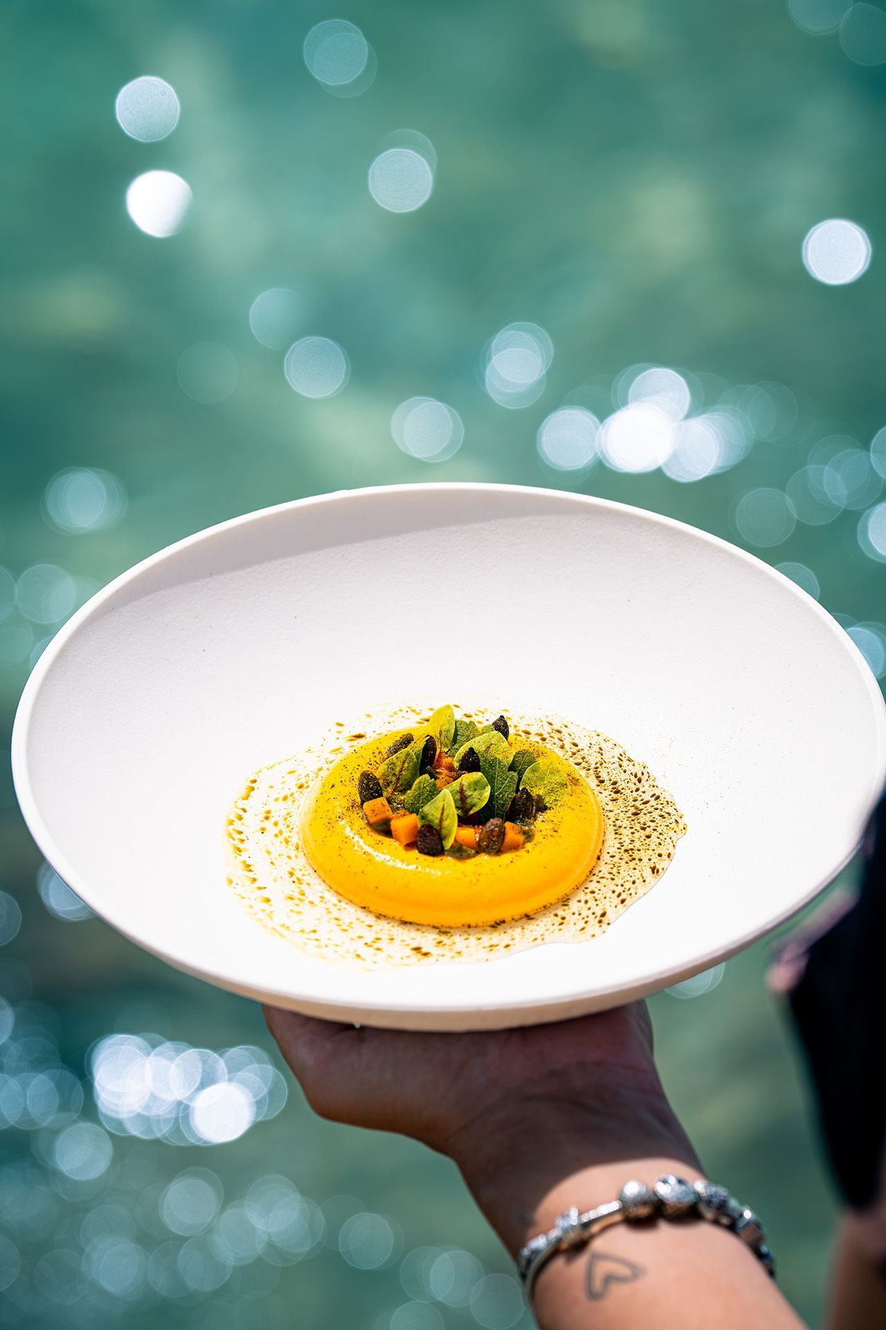A hand holding a white plate with a yellow circular dish topped with green and orange bits, set by water.