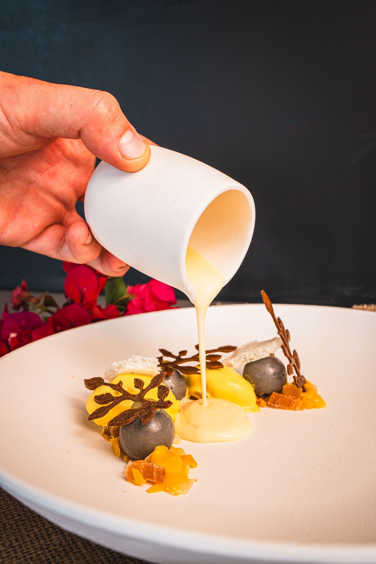 Hand pouring yellow sauce onto a plated dessert with dark spheres, caramel and chocolate decorations.