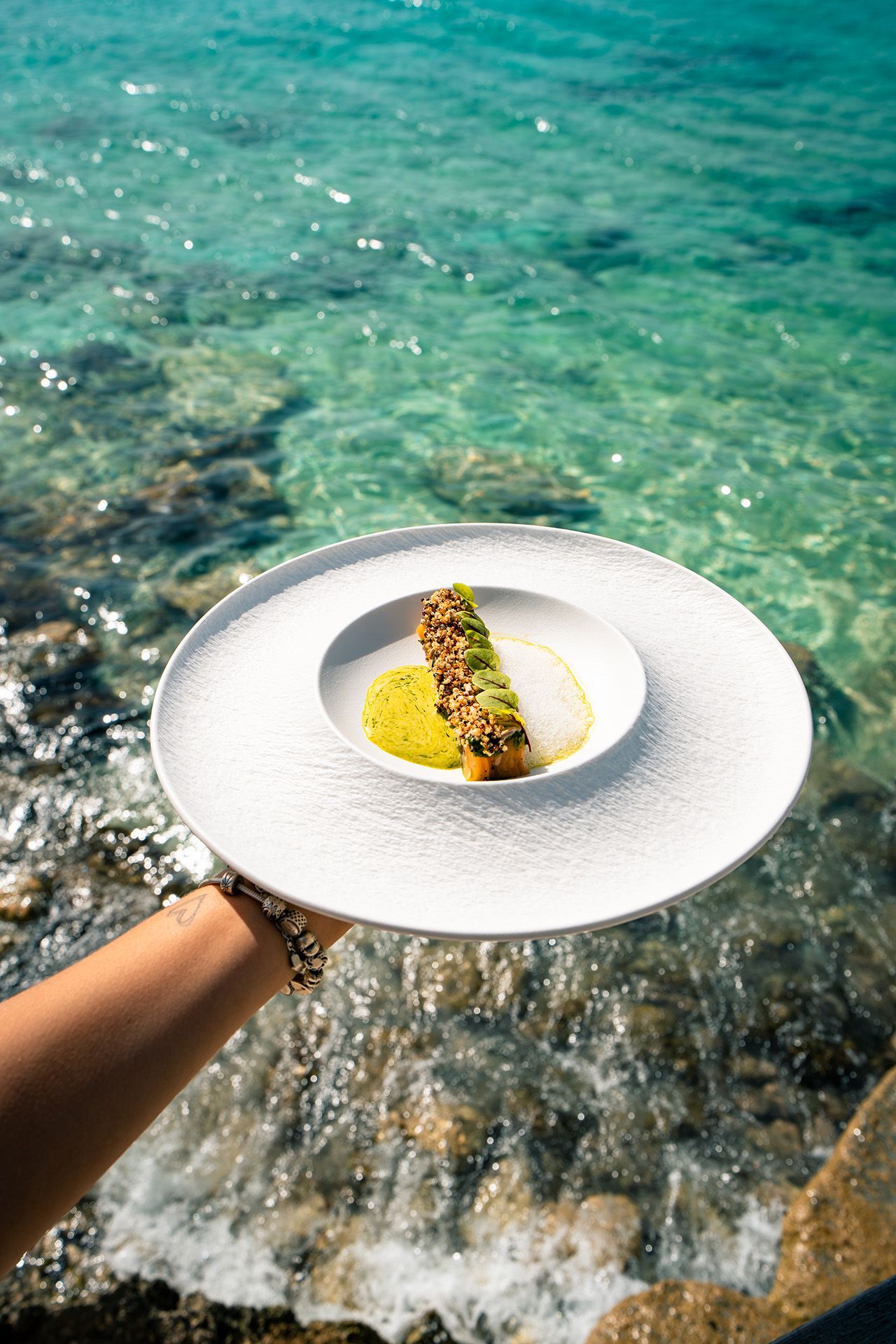 Hand holding a white plate with food, teal ocean in the background.