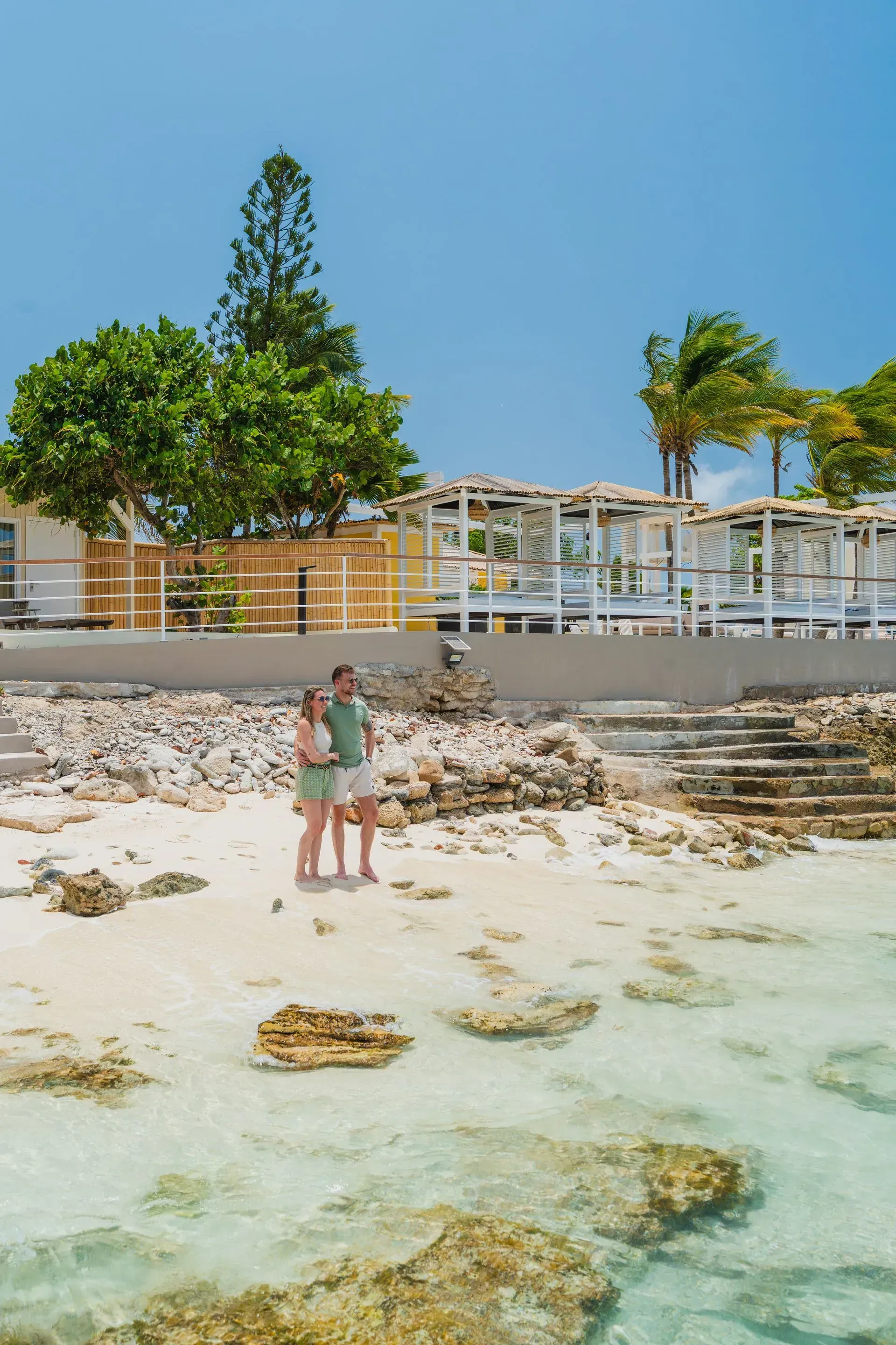 Couple stands on a sandy beach with clear water, near a white structure. Blue sky and green trees in the background.