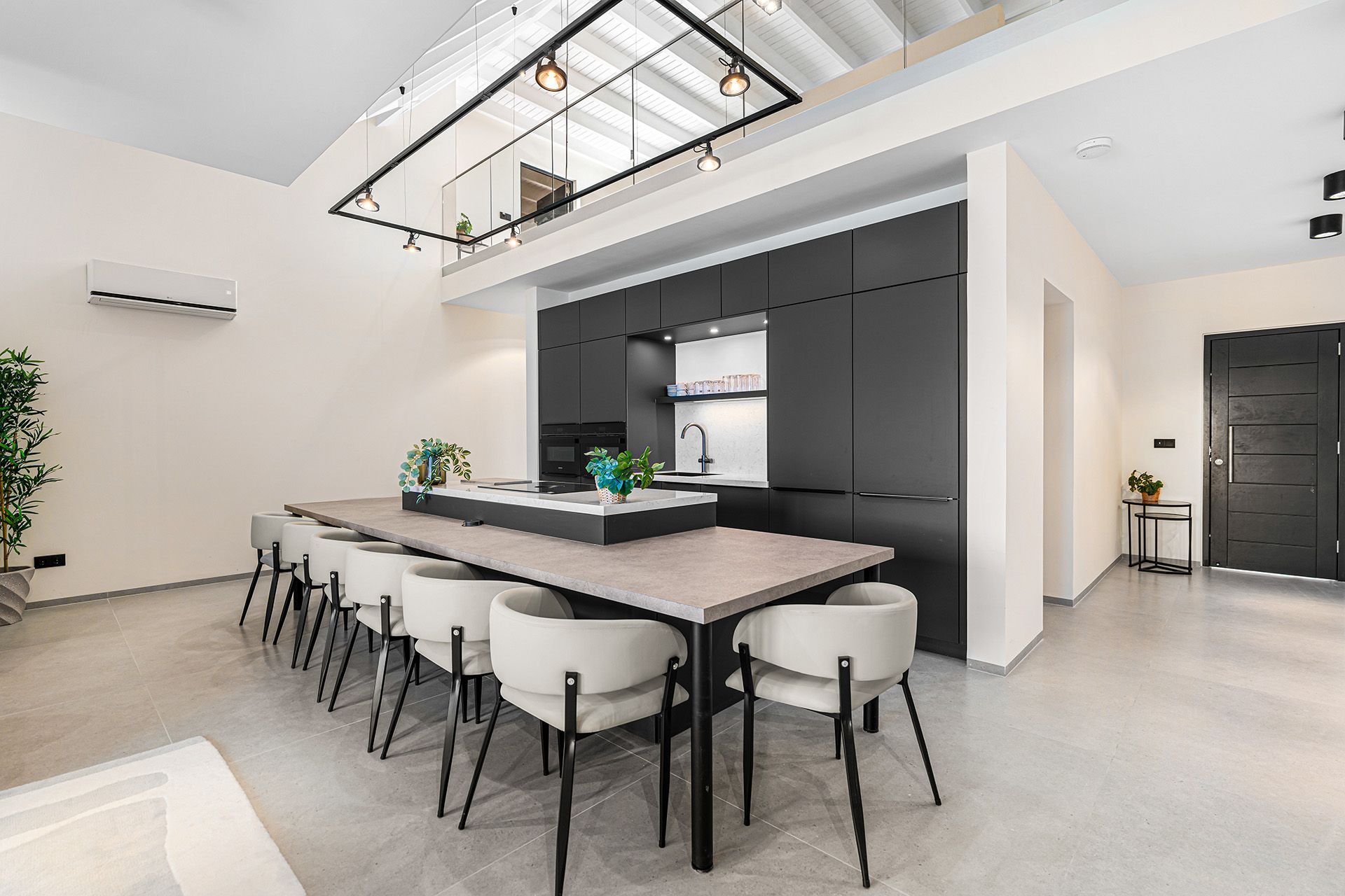 Modern kitchen with long dining table and black cabinets. Light-colored chairs and concrete floors.