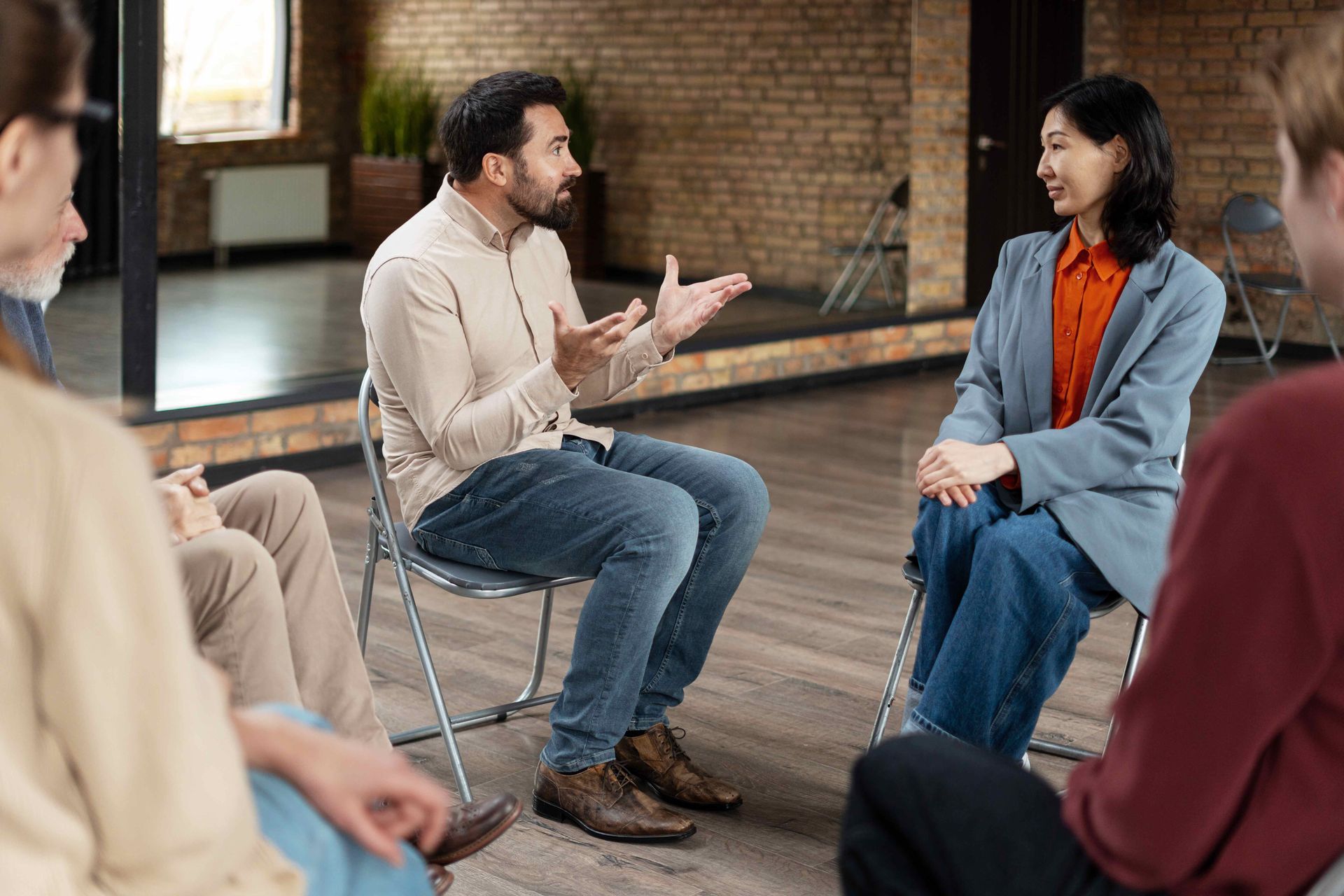 People in chairs in a circle, one person speaking, others listening in a brick-walled room.