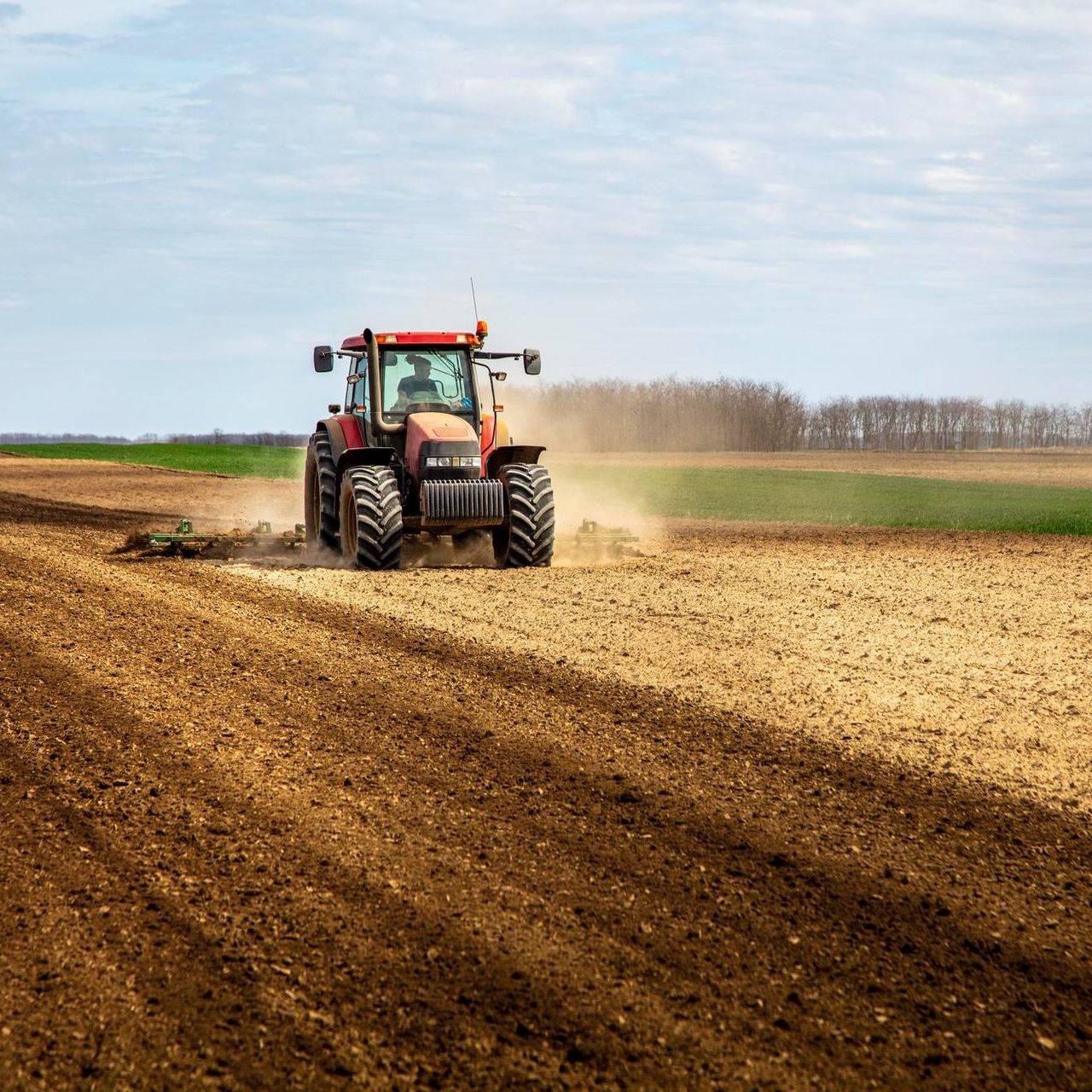 Red tractor plowing a brown field, kicking up dust, with green fields and trees in the background under a blue sky.