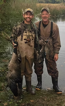 Two men in hunting gear stand by water, holding ducks they caught.