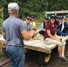 Man speaking to a group seated on a wagon in a farm setting. People listening attentively.
