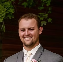 Man in a gray suit, smiling, with a small beard, and a light-colored tie, outdoors.