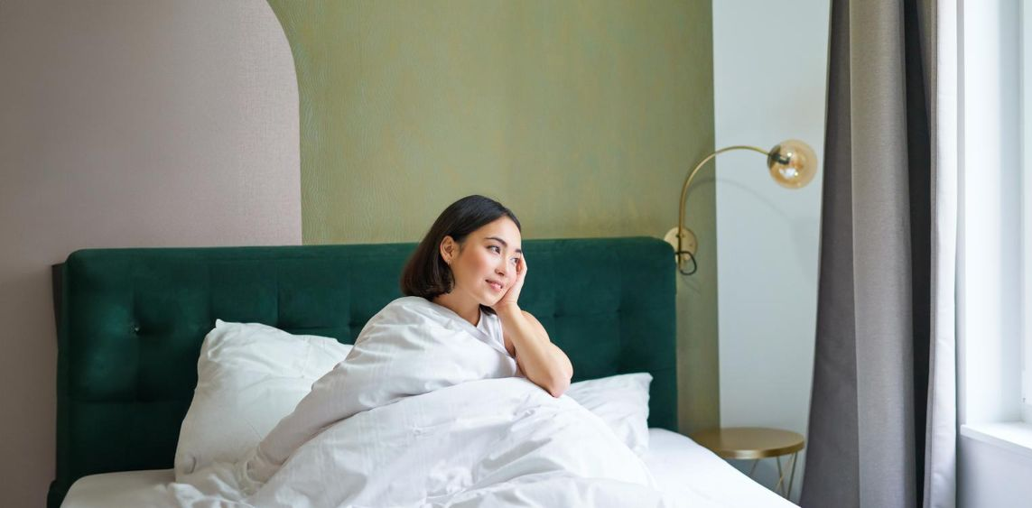 Woman sits up in bed, looking out the window.  Emerald green headboard, white bedding. Light green walls.