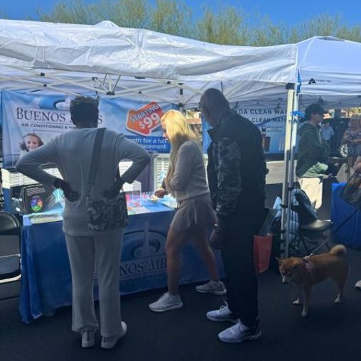 People at a market booth, looking at display. A dog on a leash, a tent overhead.