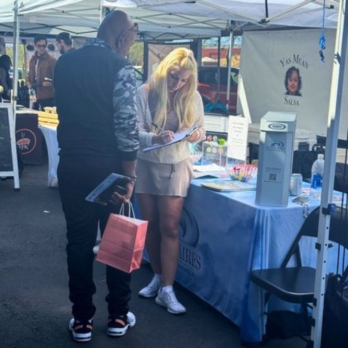 A woman writes at a table under a market tent; a person stands facing her.