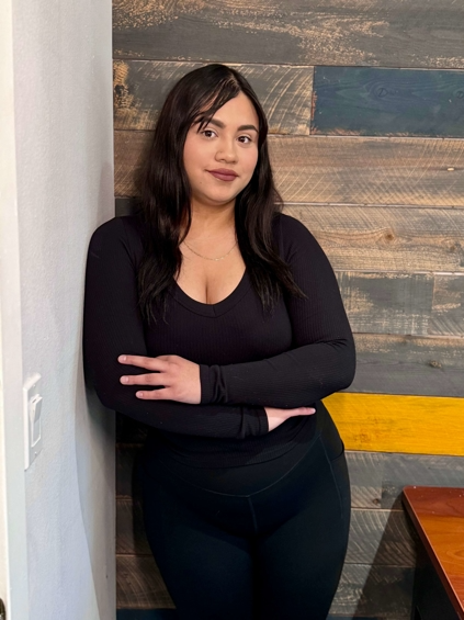 Woman in black long-sleeved shirt and leggings leans against a wood-paneled wall, arms crossed.