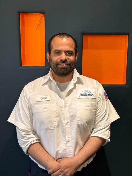Man in white work shirt, arms crossed, standing in front of blue wall with orange rectangular cutouts.