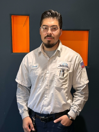 Man wearing work uniform, glasses, stands in front of a blue wall with orange rectangular cutouts.