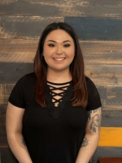 Woman with brown hair and black shirt smiles in front of a wood-paneled wall.