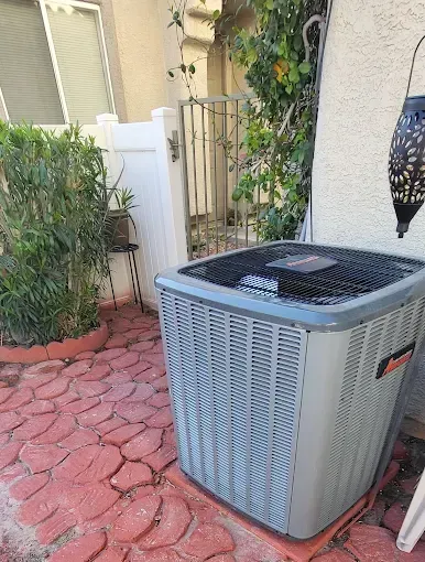 Air conditioning unit on a red brick patio with white fence and greenery in the background.