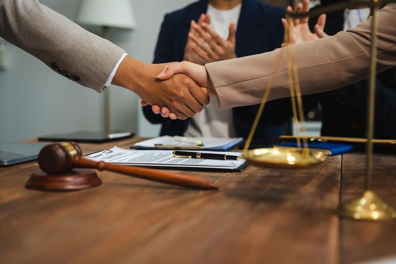 Two people shaking hands at a table, scales of justice and gavel present, others applaud in the background.