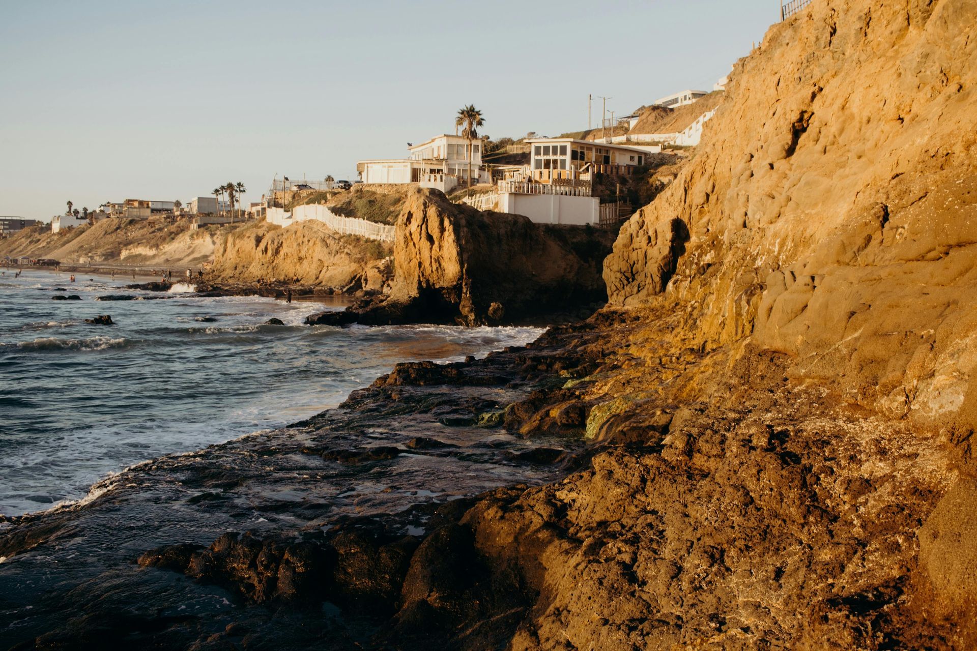 Rocky coastline with buildings on a cliff at sunset. Golden light illuminates the rocks and water.