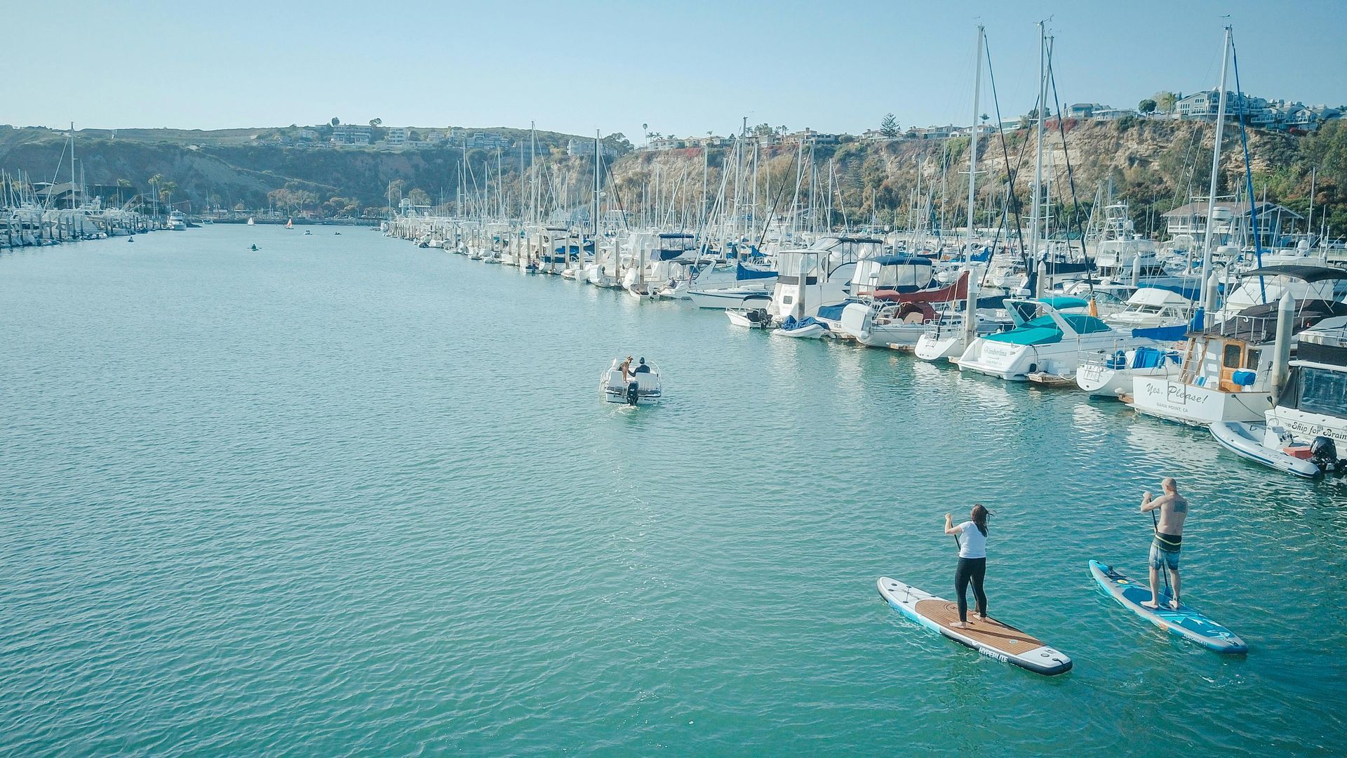 Two people paddleboard on a turquoise harbor near docked sailboats. Hillside in background.