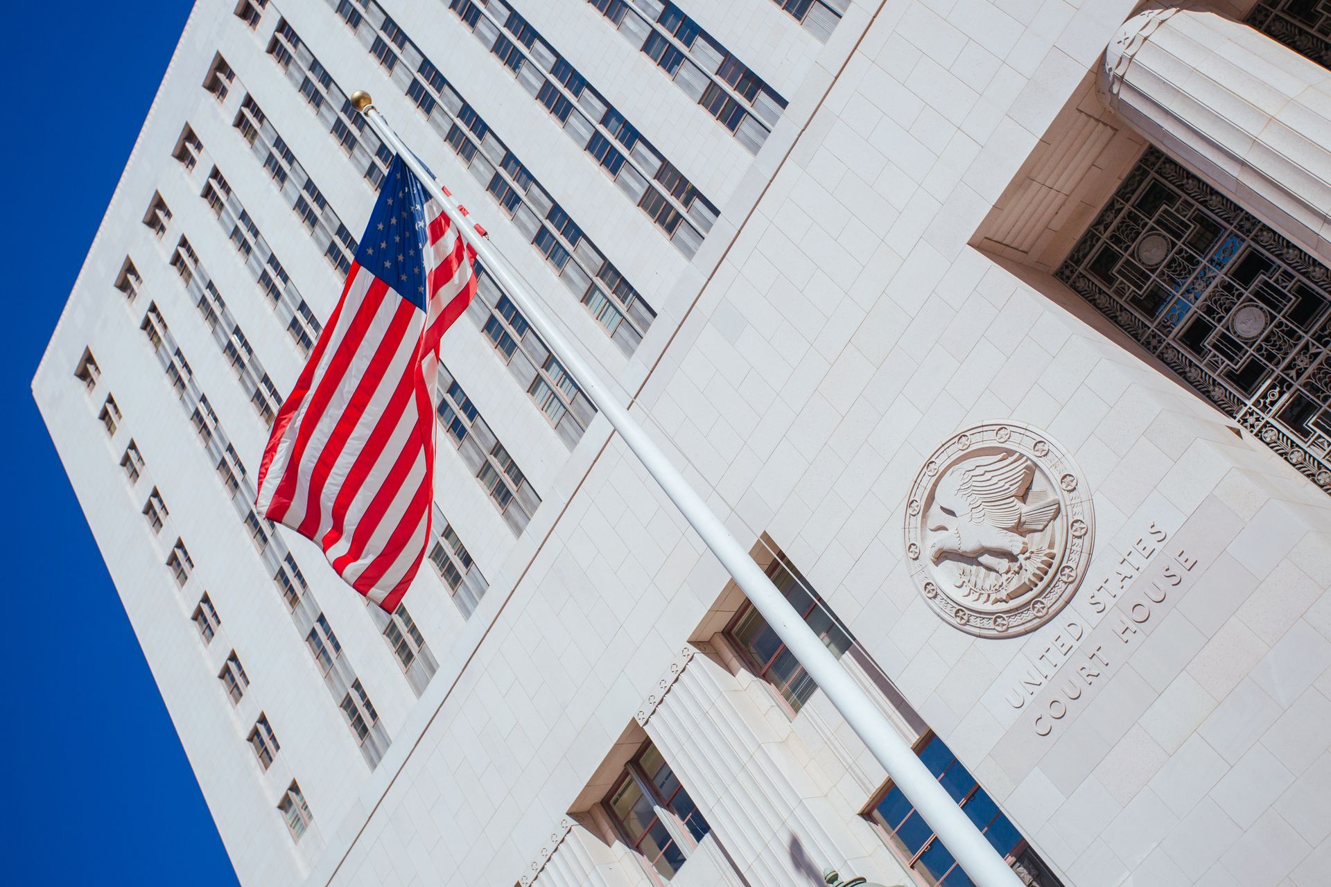 American flag at half-staff on a flagpole outside a white stone building with windows and a carved emblem.