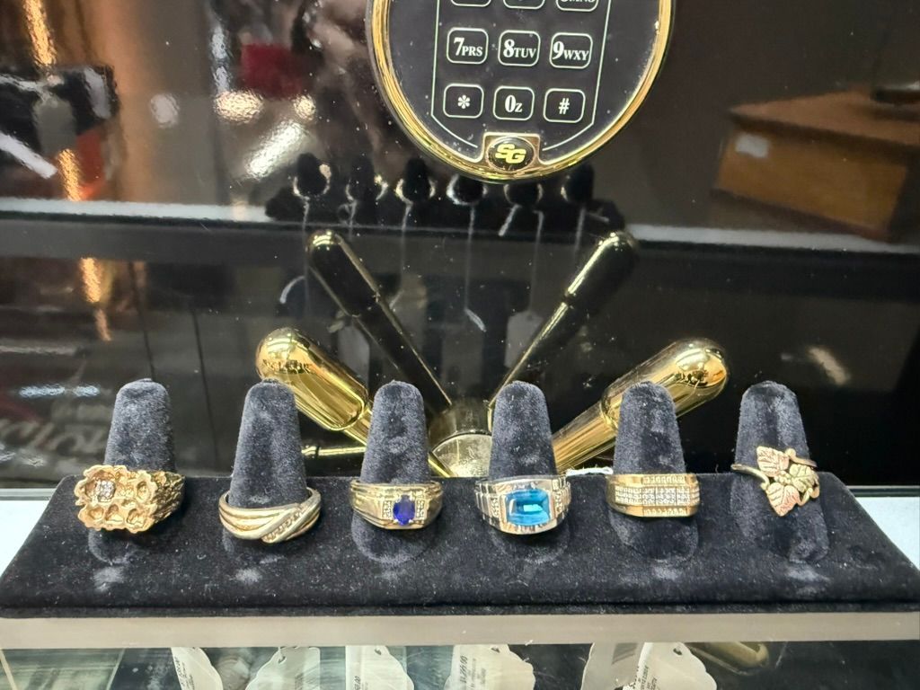 Six gold rings with various gemstones arranged on a black display stand inside a jewelry store showcase.