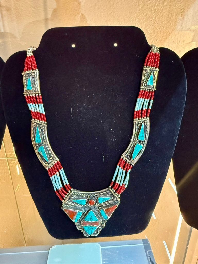 An ornate necklace with turquoise and red stone inlays, featuring beaded strands on a black display bust.