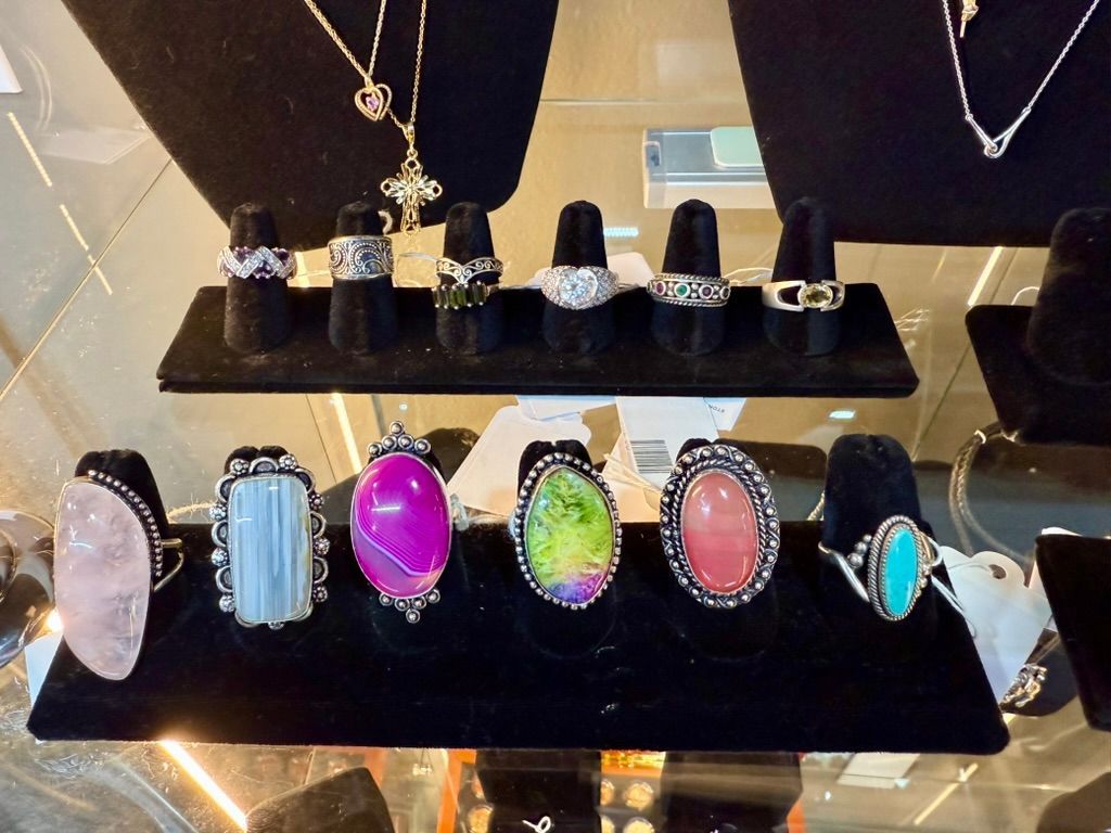 A display case with two rows of various rings featuring colorful gemstones and metal bands on black velvet stands.