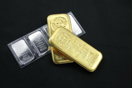 Two gold bars resting on a black surface next to small, rectangular silver bullion bars in clear plastic packaging.