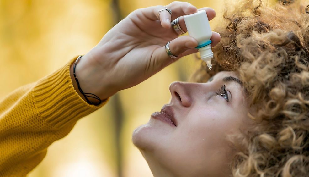 Woman tilting head back, using eye drops outdoors.