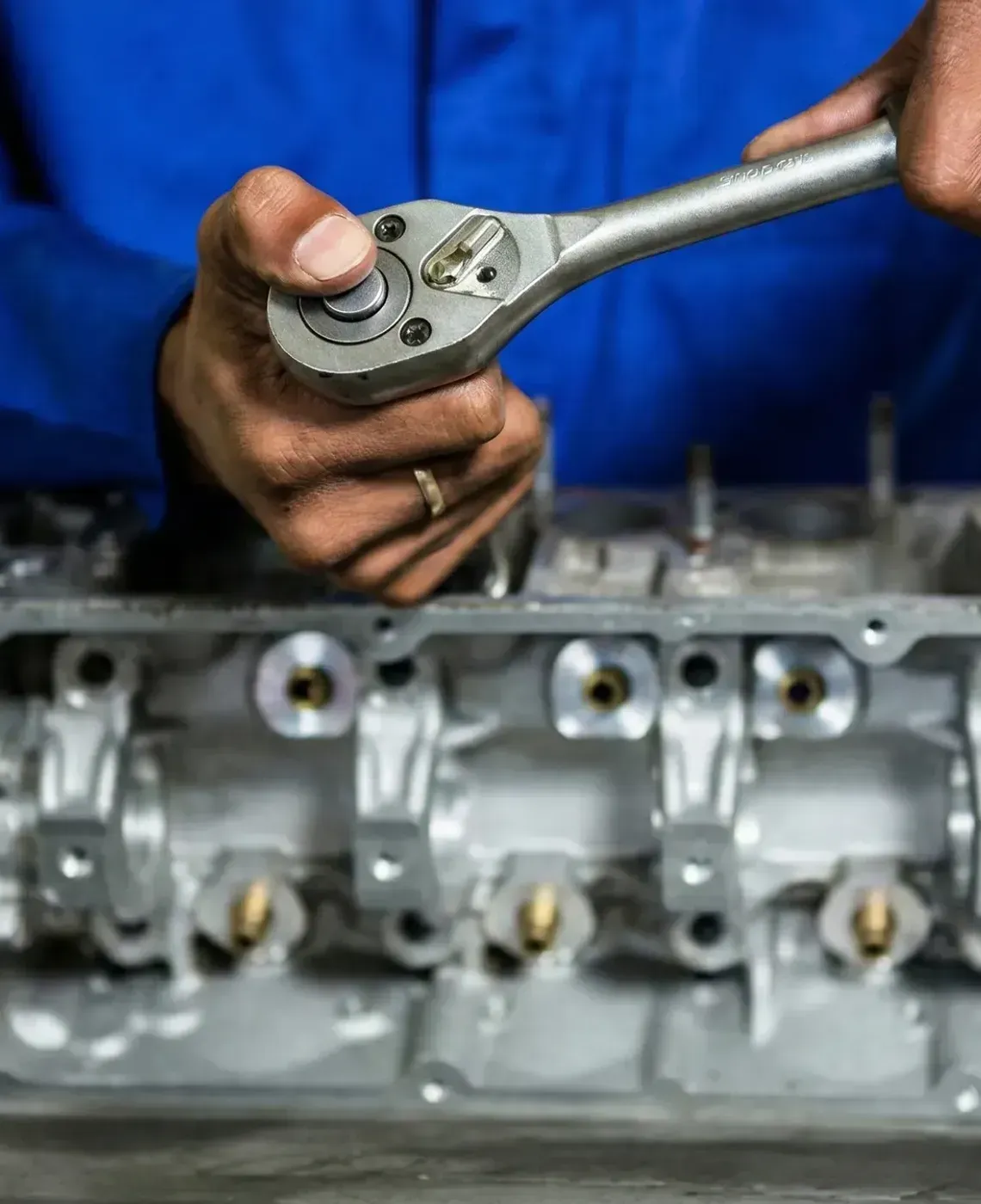 Mechanic using a ratchet to tighten bolts on a metallic engine cylinder head in a workshop.