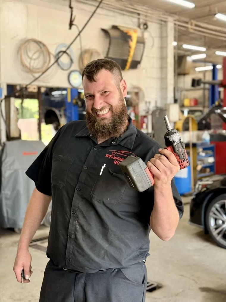 A smiling mechanic in a black uniform stands in an auto shop holding a power drill.