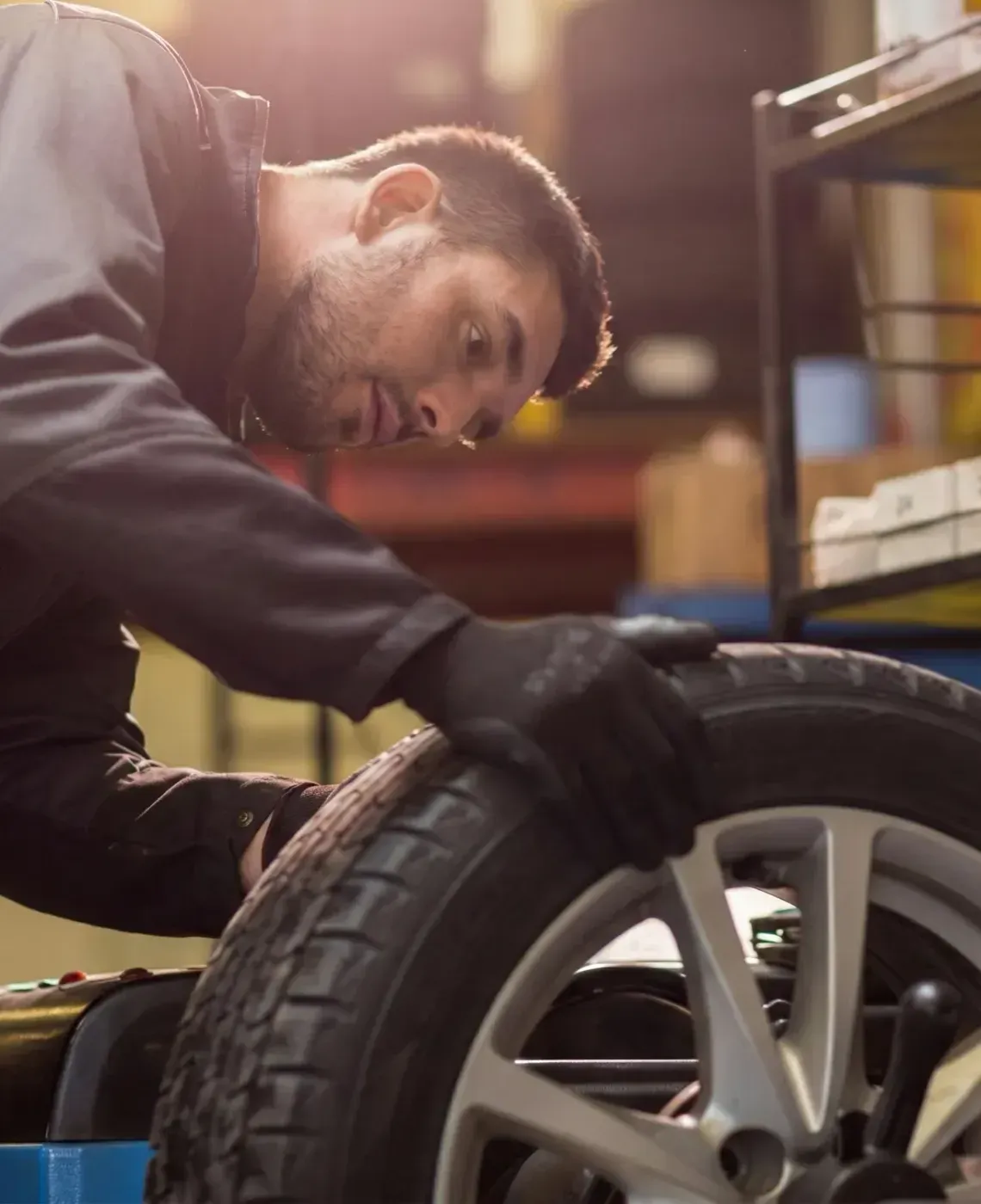 A technician in a gray uniform and black gloves examines a car tire mounted on a wheel balancing machine in a workshop.